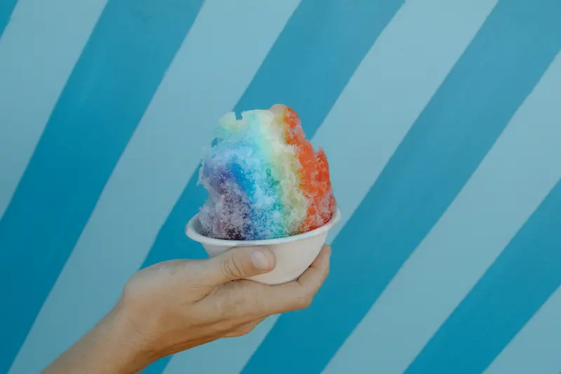 A rainbow shave ice held up against the truck’s blue stripes